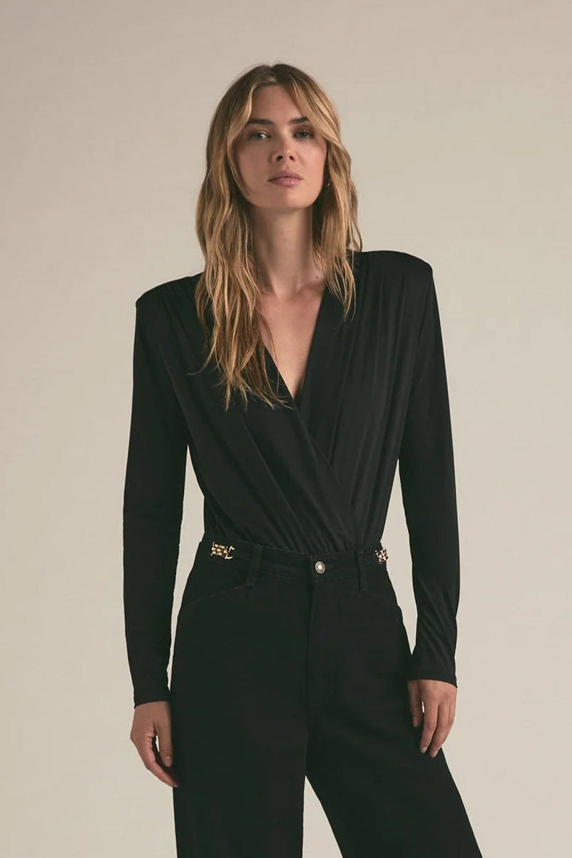 A woman modeling a black long-sleeve wrap-style jumpsuit with a defined waist and wide-leg trousers, standing against a plain beige studio background.